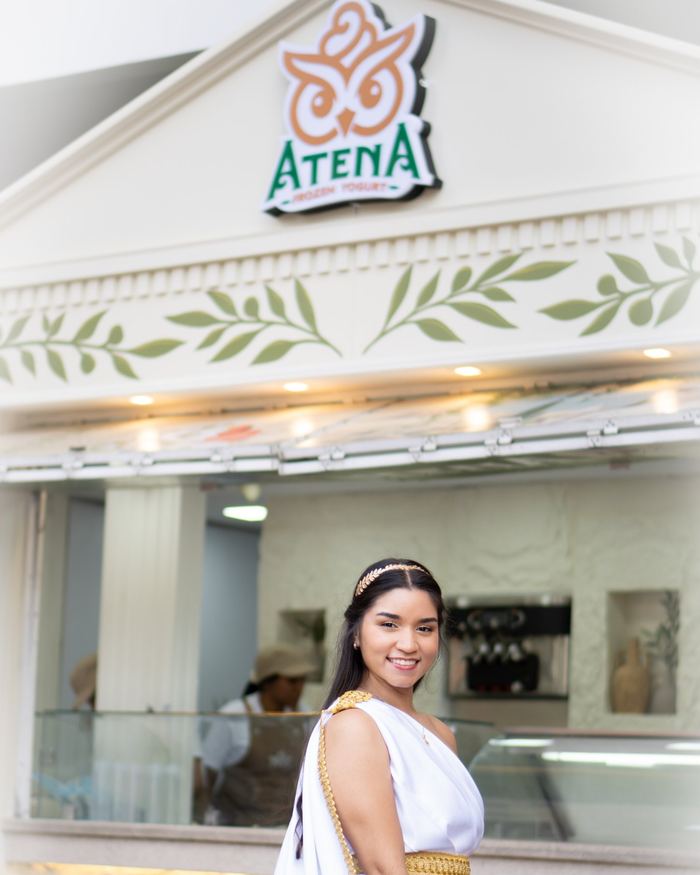 Joven vestida de diosa griega frente a la tienda de Atena, bajo el frontón con el logo
